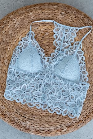 The Eye Of The Sun Padded Bralette in gray blue, featuring intricate floral lace, is displayed flat on a round woven wicker surface.