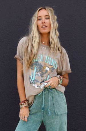 A woman with long, wavy blonde hair stands against a dark wall in a boho-chic look—she confidently wears an oversized Fly Free Graphic Tee - Mocha with an eagle print, paired with green drawstring pants and colorful beaded bracelets.
