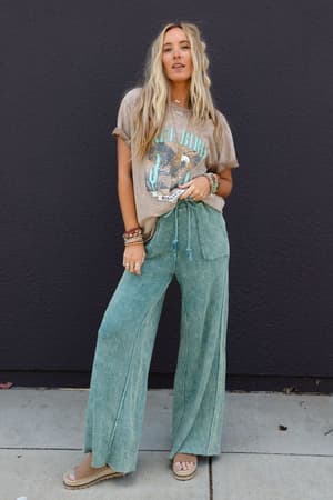 A woman with long blonde hair stands before a black wall, dressed in an oversized Fly Free Graphic Tee - Mocha, loose green pants, sandals, and layered bracelets, exuding a relaxed boho-chic vibe with one hand in her pocket.