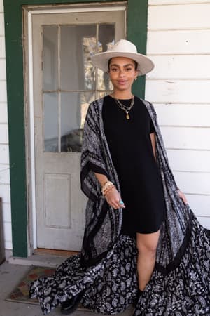 A woman stands on a porch in front of a white door, smiling slightly at the camera. She wears a wide-brimmed hat, black dress, layered necklaces, and the Paisley Tapestry Free Flow Duster Kimono - Black draping to the ground.