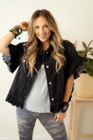 A smiling woman with long wavy hair wears the Scottie Ruffle Denim Jacket - Black over a gray tee and camouflage leggings. She poses indoors with bracelets, next to a wooden stand topped with a potted plant.