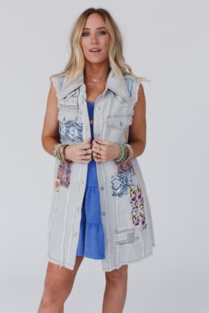 A woman with long blonde hair wears the One Of A Kind Sleeveless Denim Dress Vest - Denim Multi over a blue dress, accessorized with layered bracelets, as she stands against a plain white background.