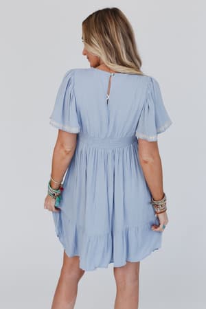 A woman with long blonde hair faces away, wearing the Magdaline Mini Dress - Blue, a light blue mini dress with short sleeves, ruffled hem, and lace trim. She accessorizes with colorful bracelets against a plain white background.