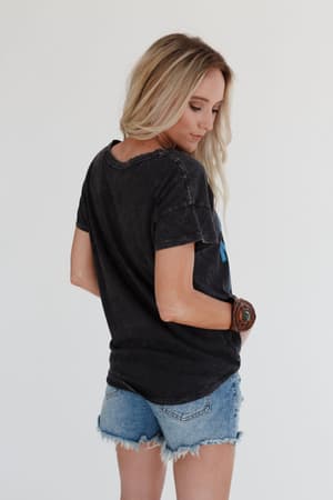 A woman with long blonde hair stands against a white background, wearing the Thunder Mama Vintage Graphic Tee in charcoal, distressed denim shorts, and a large brown bracelet, facing away and looking over her left shoulder.