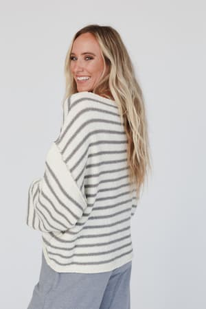 A woman with long blonde hair smiles over her shoulder in the All My Stripes Oversized Sweater - Ivory Gray, styled with light blue pants against a plain white background.