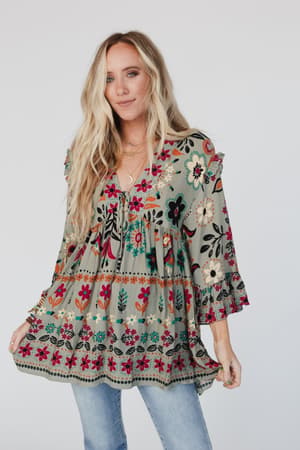 A woman with long blonde hair wears the Leona Floral Print Tunic in sage, featuring red, black, and cream patterns and a v-neckline, paired with light blue jeans. She stands against a plain white background.