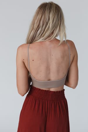 A woman with long blonde hair is seen from behind, wearing the Dora Low Back Seamless Bralette in Mocha and high-waisted rust-colored pants against a plain light gray background.