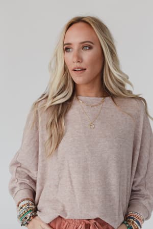 A woman with long blonde hair wears The Nest Fly With Me Batwing Sleeve Knit Sweater in Sand, layered gold necklaces, and beaded bracelets. She looks to the side with a neutral expression against a plain light background.