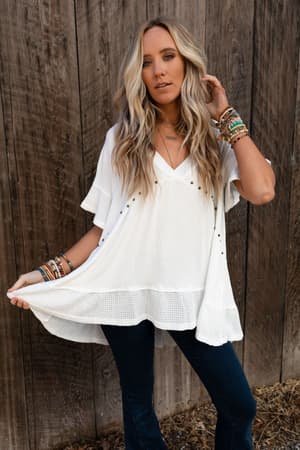 A woman with long, wavy blonde hair stands by a wooden fence wearing the More Than Meets The Eye Tunic Top in ivory, paired with dark jeans and multiple bracelets. She holds out her shirt with one hand and touches her hair with the other.