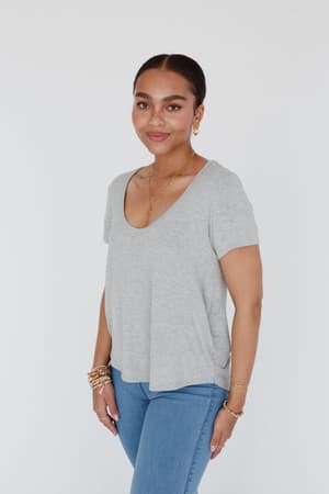 A woman stands against a white background, smiling gently. She wears the Scoop Neck Bralette Tee - Gray with blue jeans and gold jewelry, her hair pulled back.