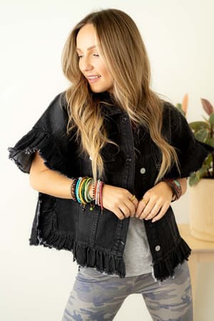 A woman with long, wavy blonde hair smiles and looks down, wearing the Scottie Ruffle Denim Jacket - Black over a grey tee and camo leggings. She shows off colorful bracelets on both wrists. A plant sits in the background.