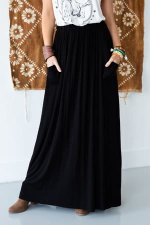 Wearing The Perfect Pocketed Maxi Skirt - Black with pockets, a white graphic sleeveless top, brown boots, and colorful bracelets, someone stands in front of a brown and white patterned hanging textile.
