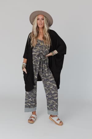A woman poses against a plain background wearing a gray wide-brim hat, camo-print jumpsuit, black kimono cardigan, white sandals, and colorful bracelets. Her look completes perfectly with The Nest Boho Essential Hooded Ribbed Sweater in Black.