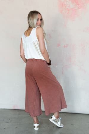 A woman with long blonde hair stands sideways, looking over her shoulder. She wears a white sleeveless top, Relaxing Robin Wide Leg Pant in Red Bean, and white sneakers, set against a light wall with pink accents.