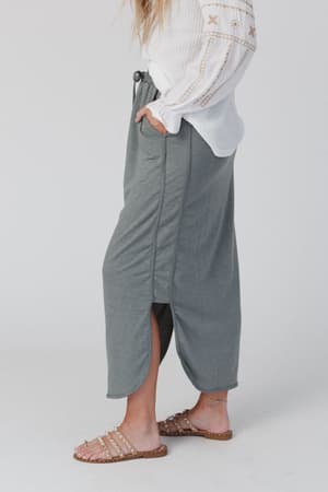 A person is seen from the waist down wearing The Nest So Comfy Drawstring Maxi Skirt in light olive, styled with a white textured long-sleeve top and tan sandals with gold studs, all set against a plain background.