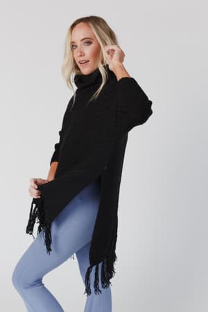 A blonde woman wears the Paloma Poncho Sweater in black with fringed edges and light blue leggings, standing sideways and looking at the camera against a plain white background.