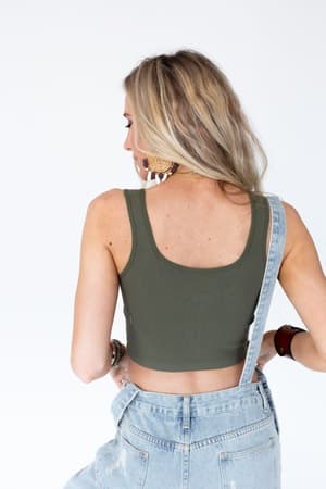 A woman with long blonde hair, wearing the Hook It Up Ribbed Brami in olive and light blue denim overalls with one strap off her shoulder, stands facing away against a plain white background.