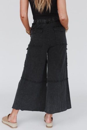 A woman stands facing away, wearing the Game On Cargo Wide Leg Pant in black with a ruffled waistband, paired with a black top, bracelets, and beige espadrille sandals.