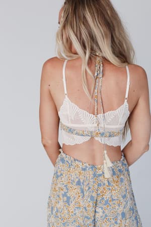 A woman with long blonde hair is seen from the back wearing the Butterfly Beauty Bralette - Ivory and high-waisted blue patterned pants with yellow and white designs. A tasseled string trails down her back.