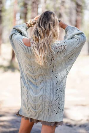 A woman with wavy blonde hair stands outdoors, facing away, wearing the Golden Hour Bubble Sleeve Sweater Dress in sage. She holds her hands to her head and has bracelets on her wrist.