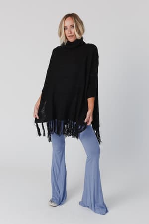 A woman with light blonde, wavy hair wears the Paloma Poncho Sweater - Black, featuring a fringed oversized silhouette, paired with light blue flared pants against a plain background.