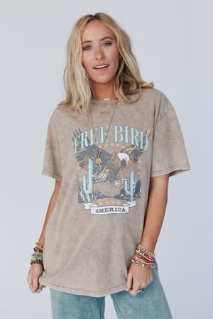 A woman with long blonde hair wears the Fly Free Graphic Tee - Mocha, an oversized tee with an eagle and cactus design, styled with blue jeans and colorful bracelets, standing against a plain light background.