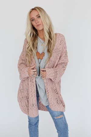 A woman with long, wavy blonde hair wears the Cara Oversized Cardigan in pink over a gray top and ripped blue jeans, accessorized with layered necklaces and rings—an ideal transitional layering piece against a simple white background.