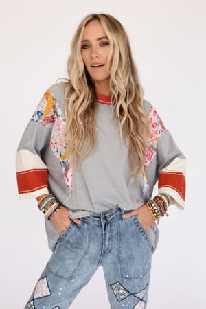 A woman with long, wavy blonde hair wears the Britt Patchwork Top in Heather Gray, featuring 3/4-length colorful patchwork sleeves, paired with blue jeans with patchwork details. She stands hands-in-pockets, accessorized with bracelets against a plain background.