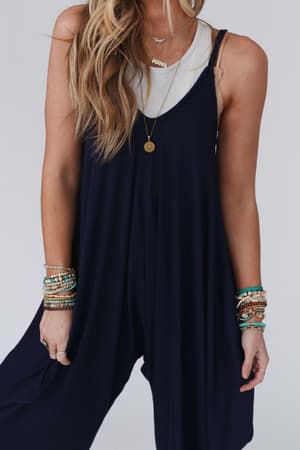 A woman wears The Perfect Harem Jumpsuit - Navy over a white tank top, paired with layered necklaces and colorful beaded bracelets. Her long, wavy blonde hair drapes over one shoulder.