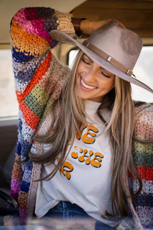 A smiling woman with long hair wears the Wrapped up in Color Knit Cardigan - Cream, a wide-brimmed hat, and a white graphic tee. She sits in a car, tilts her head, and lifts her arm while enjoying a joyful moment.