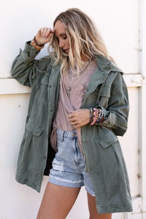 A woman with long blonde hair wears the Layers On Layers Perfect Boho Parka in olive, along with a taupe tee, distressed denim shorts, and layered jewelry. She stands against a white wall, looking down and touching her hair.