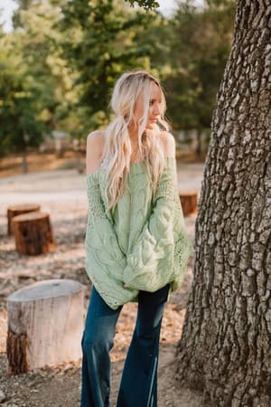 A woman with long, wavy blonde hair stands outdoors near a tree, wearing the Golden Hour Bubble Sleeve Sweater Dress in light green, paired with dark blue flared jeans. Tree stumps and greenery are visible in the background.