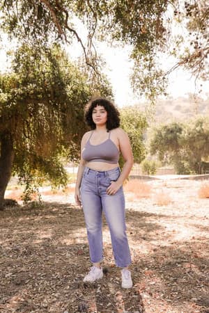 A woman with curly hair stands outdoors on a sunny dirt path surrounded by trees, wearing the Tattoo Bralette in gray, high-waisted blue jeans, and white sneakers.