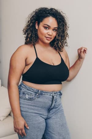 A woman with curly hair wears the Tattoo Bralette - Black with high-waisted blue jeans, standing indoors and smiling confidently at the camera.