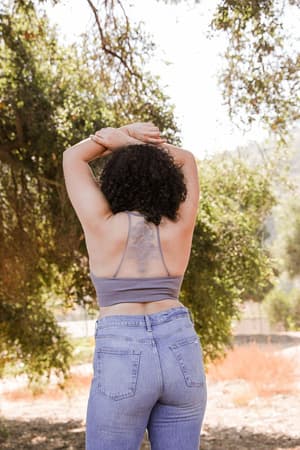 Wearing the Tattoo Bralette - Gray under a gray tank top, a curly-haired person stands outdoors with their back to the camera and arms raised, surrounded by trees and sunlight.