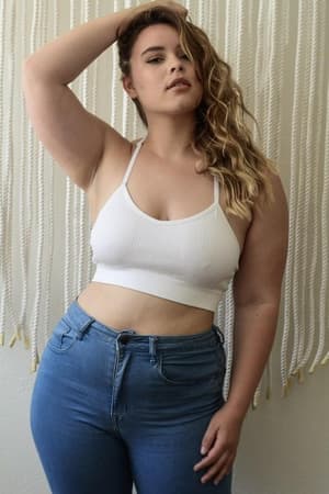 A woman with wavy blonde hair poses with one hand in her hair, wearing the Tattoo Bralette - White and blue jeans, standing before a wall decorated with hanging white ropes.