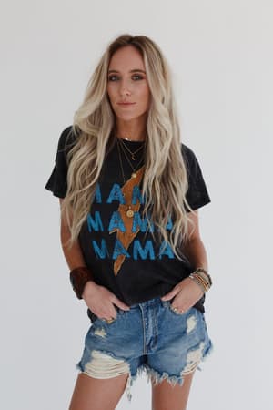 A woman with long, wavy blonde hair wears the Thunder Mama Vintage Graphic Tee in charcoal, featuring distressed "MAMA" lettering with a yellow lightning bolt, styled with denim shorts and layered necklaces against a plain white background.