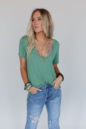 A woman with long blonde hair wears the Scoop Neck Bralette Tee in Sage over a lace bralette, paired with blue ripped jeans, layered necklaces, and beaded bracelets, standing against a plain light background.