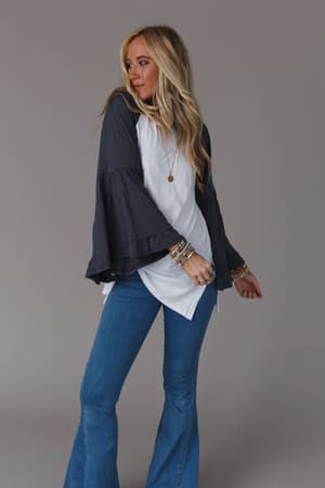 A woman with long blonde hair wears the Bell Sleeve Baseball Tee in Charcoal and blue flared jeans, accessorized with colorful bracelets. She stands against a plain gray background, looking to the side in a relaxed pose.