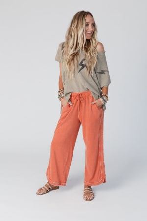 A woman with long wavy blonde hair smiles against a plain background, wearing the Struck By You Dolman Sleeve Sweater Top in mocha with black lightning bolt prints, loose orange drawstring pants, stacked bracelets, and strappy sandals.