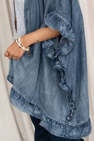 A person wearing the Robbie Denim Jacket - Blue, styled oversized with ruffle trim, a white shirt, dark pants, and bracelets, is photographed from the side against a neutral background.