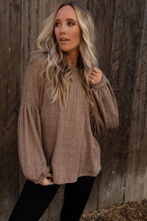 A woman with long, wavy blonde hair stands before a wooden wall, wearing The Nest Sweet Heart Balloon Sleeve Knit Sweater in Mocha and black pants. She gazes to the side, her sweater's wide sleeves adding a stylish flair to her casual look.