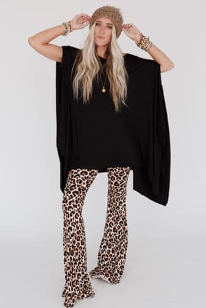 A woman wearing The Wren Tunic - Black, leopard print flared pants, a brown knit beanie, and colorful beaded bracelets poses with her hands on her hat against a plain white background.