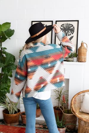 A woman with long blonde hair, wearing a wide-brimmed black hat, blue jeans, a white shirt, and the Anika Mixed Pattern Jacket - Teal, stands with her back to the camera in a cozy, plant-filled room.