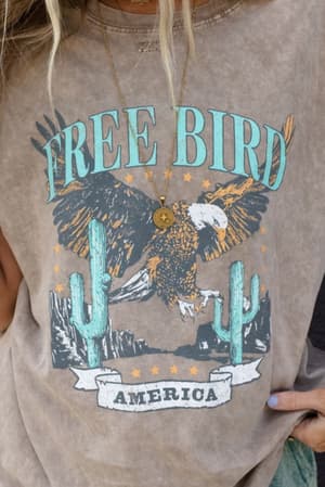 A person wears the Fly Free Graphic Tee - Mocha, an oversized faded brown shirt with an eagle soaring among cacti, stars, and mountains. “FREE BIRD” and “AMERICA” are printed on it for a boho-chic vibe, styled with a necklace.