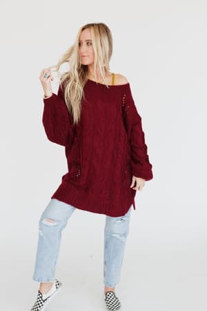 A woman with long blonde hair wears the Golden Hour Bubble Sleeve Sweater Dress in burgundy, standing against a plain white background as she holds a strand of her hair and gazes to the side.