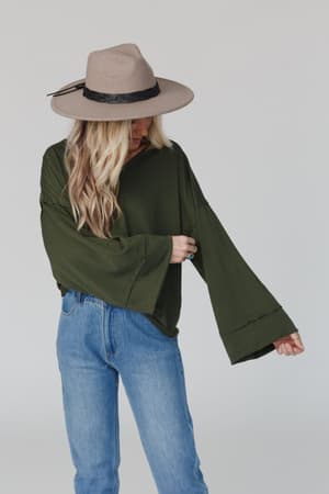 A person with long blonde hair, wearing a wide-brimmed tan hat, The Nest Love It V Neckline Sweater Top in olive, and blue jeans stands against a plain light background, looking down with one arm slightly raised.