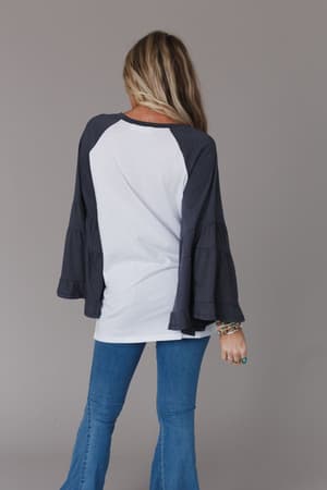 A woman with long blonde hair faces away, wearing the Bell Sleeve Baseball Tee - Charcoal with dark ruffled sleeves and blue jeans. She has bracelets on her left wrist and stands against a plain gray background.