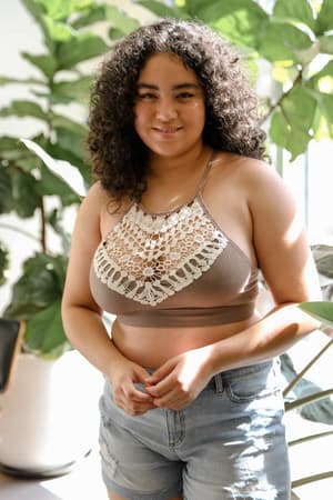 A woman with curly hair smiles while wearing the Crochet High Neck Bralette - Mocha and denim shorts, standing indoors near green potted plants in natural sunlight.