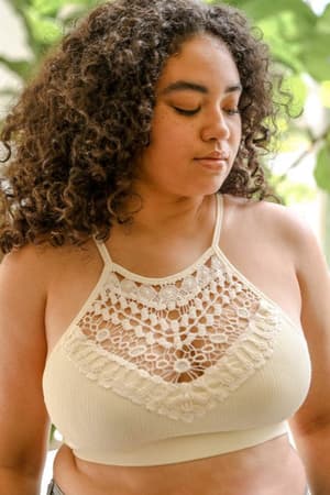 A woman with curly hair wears the Crochet High Neck Bralette in Ivory outdoors, looking down peacefully. Green foliage in the background highlights this ideal layering piece for a relaxed summer vibe.